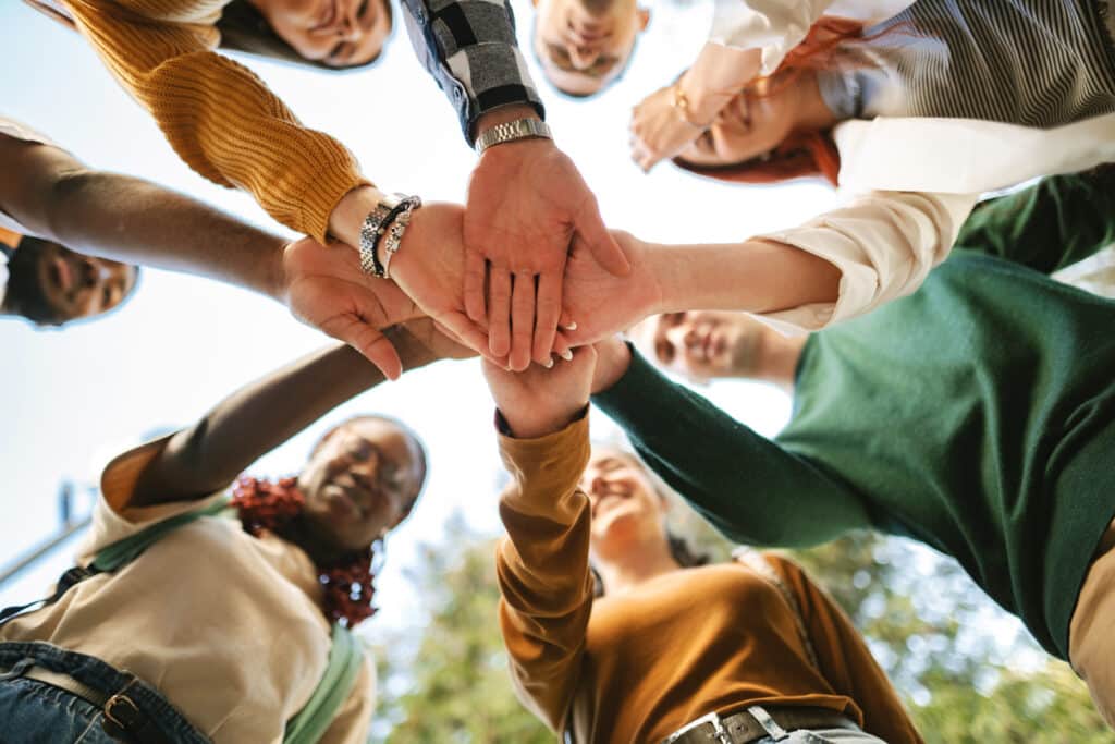 Students from various backgrounds join hands in a supportive circle, celebrating their bond and commitment to teamwork in an outdoor setting on campus.