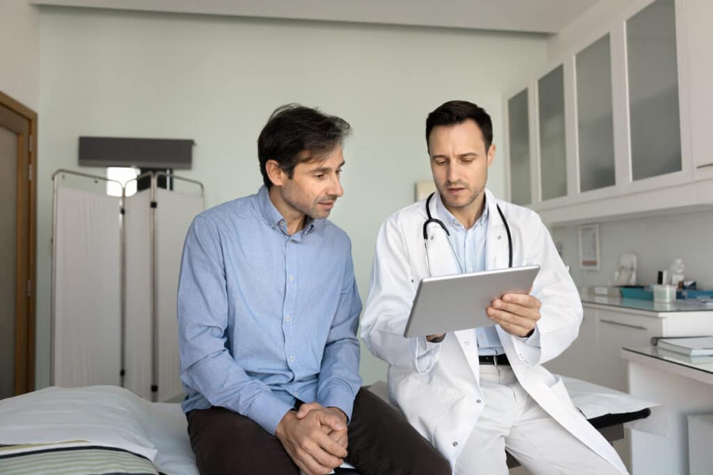 Serious doctor showing electronic checkup results to patient, using tablet computer, talking to man, explaining medical report, diagnosis after examination in clinic office